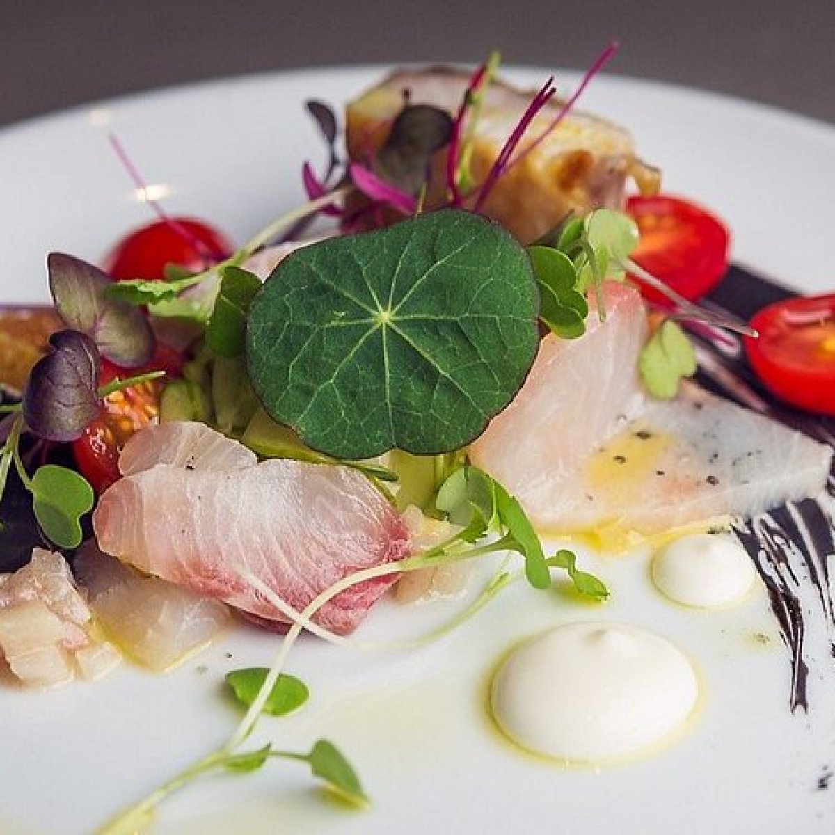 Gourmet dish with fish, greens, cherry tomatoes, sauces on a white plate.